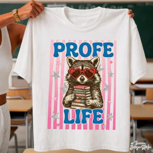 May include: White t-shirt featuring a raccoon wearing heart-shaped sunglasses and holding books. The text "PROFE LIFE" is in blue, with pink stripes and silver stars in the background. The shirt is held up by a person.