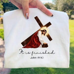 May include: White sweatshirt featuring a religious image of Jesus kneeling before a cross. The image includes a halo and a red robe. The text "It is finished" and "John 19:30" are printed below the image.