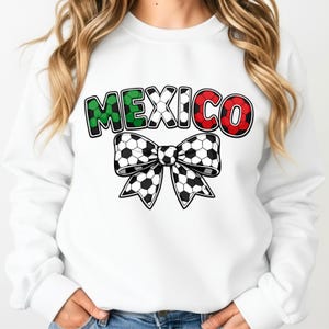 Op de afbeelding: Witte sweatshirt met het woord "MEXICO" in groen en rood, met een voetbalstrikontwerp. De letters zijn zwart omlijnd. De strik is zwart-wit met een voetbalpatroon.