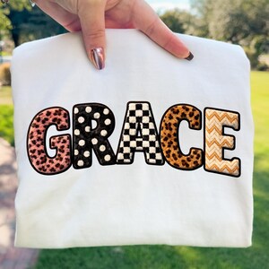 May include: A white sweatshirt featuring the word "GRACE" in bold, patterned letters. The letters are decorated with leopard print, polka dots, and a chequered design. The background is a blurred view of green and blue.
