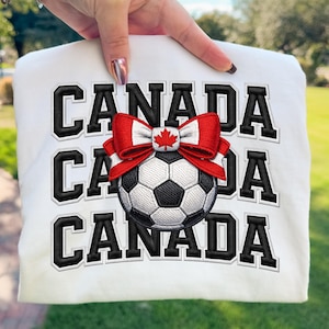 May include: White sweatshirt with the word "CANADA" in black block letters. A red and white bow with a maple leaf emblem sits above a black and white soccer ball. The sweatshirt is held up by a hand.