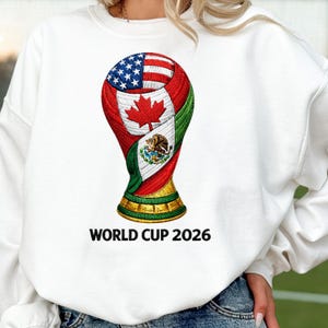 Può includere: Felpa bianca con un design a trofeo stilizzato che incorpora le bandiere di Stati Uniti, Canada e Messico. Il trofeo è rosso, bianco, verde e oro, con la scritta "WORLD CUP 2026" sotto.