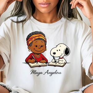 May include: White t-shirt featuring a cartoon illustration of Maya Angelou and Snoopy writing in notebooks. Maya Angelou is depicted in a red shirt and a yellow and orange head wrap. The text "Maya Angelou" is printed below the illustration.