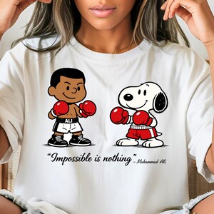 May include: White t-shirt featuring cartoon illustrations of a boxer and Snoopy wearing red boxing gloves. The phrase "Impossible is nothing" is printed below the illustrations, attributed to Muhammad Ali.