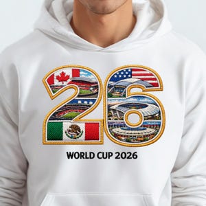 May include: White hoodie featuring an embroidered design with the number "26" filled with stadium images and flags of Canada, the United States, and Mexico. The text "WORLD CUP 2026" is printed below the number. The design has a gold border.