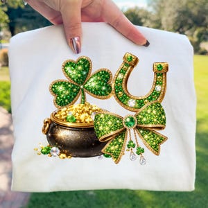 Puede incluir: Tela blanca con un diseño del Día de San Patricio. El diseño incluye un trébol verde, una herradura dorada, una olla negra llena de monedas de oro y un lazo verde con tréboles y cristales colgantes.