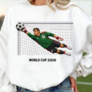 Könnte beinhalten: Weißes Sweatshirt mit einem Fußballtorwart, der nach einem Fußball greift. Der Torwart trägt ein grünes Trikot mit roten Akzenten und schwarze Shorts. Der Text "WORLD CUP 2026" ist darunter gedruckt.