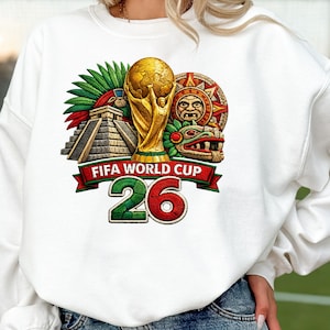 Puede incluir: Sudadera blanca con un diseño bordado colorido. El diseño incluye un trofeo dorado, una pirámide y el texto "FIFA WORLD CUP 26" en rojo y verde. La sudadera es de estilo casual.