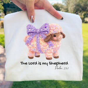 Puede incluir: Tela blanca con una colorida ilustración de una oveja con un lazo morado. La oveja tiene una apariencia esponjosa y texturizada con orejas y patas marrones. El texto debajo dice "The Lord is my Shepherd Salmo 23:1".