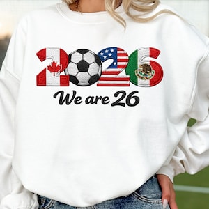Könnte beinhalten: Weißes Sweatshirt mit einem Fußball und der Zahl 26, mit den Flaggen von Kanada, den Vereinigten Staaten und Mexiko. Der Text "We are 26" ist darunter gedruckt. Das Sweatshirt ist aus weichem Material.