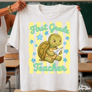 May include: White t-shirt featuring a cartoon turtle holding a paper airplane. The shirt has the words "First Grade Teacher" in a whimsical font, with blue stars and yellow stripes as design elements.