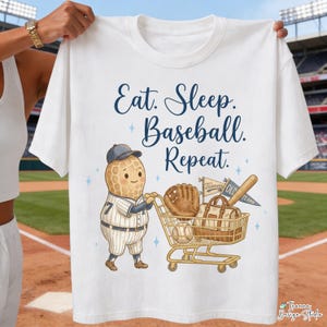 Könnte beinhalten: Weißes T-Shirt mit dem Text "Eat. Sleep. Baseball. Repeat." in Marineblau. Das Shirt zeigt eine Cartoon-Erdnussfigur, die einen Einkaufswagen mit Baseball-Ausrüstung schiebt, darunter ein Handschuh, ein Schläger und ein Ball.