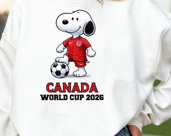 Może przedstawiać: Biała bluza z postacią z kreskówki Snoopy w czerwonym stroju piłkarskim, stojąca na piłce nożnej. Tekst "CANADA WORLD CUP 2026" jest wydrukowany poniżej w kolorze czerwonym i czarnym.