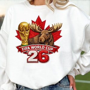 May include: White sweatshirt featuring a moose with antlers, a red maple leaf, and a gold trophy. The words "FIFA WORLD CUP 26" are displayed on a red banner. The design is a detailed graphic.
