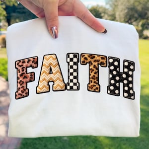 Puede incluir: Sudadera blanca con la palabra "FAITH" en letras decorativas y en negrita. Las letras tienen patrones únicos: estampado de leopardo, galones, cuadros, estampado de leopardo y lunares. La sudadera es sostenida por una mano.