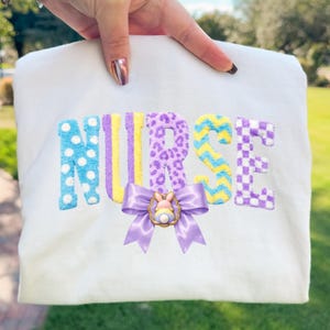 Puede incluir: Sudadera blanca con la palabra "NURSE" bordada en patrones coloridos. La "N" es azul con lunares blancos, la "U" y la "R" son amarillas y moradas, la "S" es amarilla y azul en zigzag, y la "E" es morada y blanca a cuadros. Un lazo morado con un dije de conejito en la parte inferior.