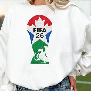 Puede incluir: Sudadera blanca con un diseño bordado colorido. El diseño incluye una hoja de arce roja, una estrella azul y el texto "FIFA 26". Un emblema verde y blanco se encuentra en la parte inferior del diseño.