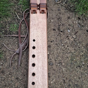Könnte beinhalten: Handgefertigte Holzflöte mit einem Federdesign, das auf das untere Ende geätzt ist. Die Flöte hat sechs Fingerlöcher und zwei braune Lederriemen. Das Instrument ist hellbraun und liegt auf einer Erdoberfläche.