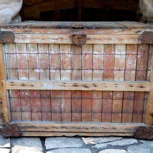 May include: A weathered wooden chest with a hinged lid and metal hardware. The front of the chest features a grid pattern with red and white accents. The chest is rectangular and sits on small, rusted feet.