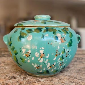 May include: A turquoise ceramic lidded bowl with a floral design. The bowl has two handles and is decorated with green, orange, and white floral patterns. The lid has a small knob and matches the bowl's color and design.
