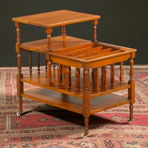 Puede incluir: Una mesa auxiliar de madera de tres niveles con acabado marrón cálido. La mesa tiene una superficie superior, un estante central con divisores y un estante inferior. Tiene patas torneadas y pequeñas ruedas.