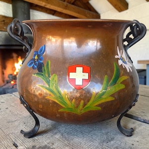 May include: A copper cauldron with black handles and legs. The cauldron features a Swiss coat of arms and floral designs in blue, white, and green. The cauldron is sitting on a wooden surface.