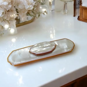 Puede incluir: Una bandeja de tocador nacarada con un jabonera a juego. La bandeja y el jabonera tienen forma rectangular con un borde marrón. El conjunto se exhibe sobre una superficie blanca, con un arreglo floral en el fondo.