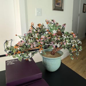 May include: A decorative bonsai tree with a brown trunk, green leaves, and pink and orange flowers. The tree is in a light blue ceramic pot. A purple box with the text "JIMMY CHOO NEW YORK" is in the foreground.