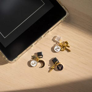 Może przedstawiać: Trzy złote zawieszki do telefonu z czarno-białymi emaliowanymi detalami. Jedna zawieszka przedstawia kokardę, druga księżyc i gwiazdę, a trzecia błyskawicę. Każda zawieszka ma inną literę: Z, J i V. Zawieszki leżą obok telefonu.