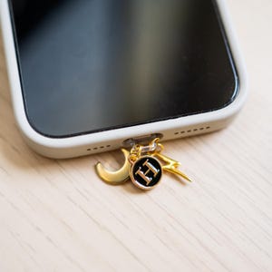 Może przedstawiać: Złoty urok do telefonu z p&oacute;łksiężycem, błyskawicą i okrągłym urokiem z literą "H" na czarnym tle. Urok jest przymocowany do białego etui na telefon.