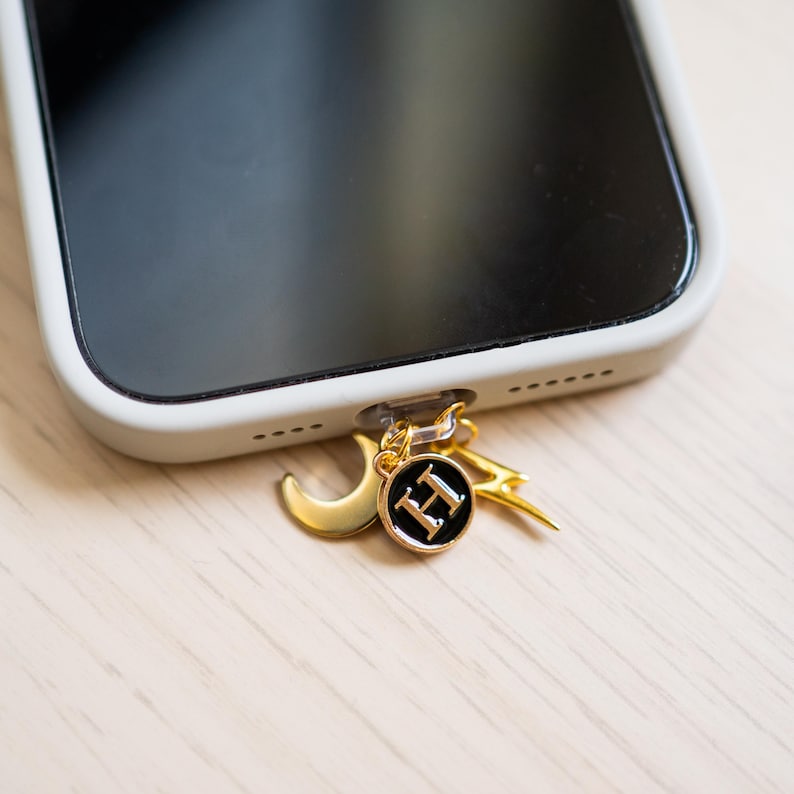 Może przedstawiać: Złoty urok do telefonu z p&oacute;łksiężycem, błyskawicą i okrągłym urokiem z literą "H" na czarnym tle. Urok jest przymocowany do białego etui na telefon.