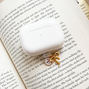 Może przedstawiać: Białe etui na AirPods ze złotym urokiem w kształcie kokardy i okrągłym urokiem z literą "Z". Etui spoczywa na otwartej książce z nadrukowanym tekstem.