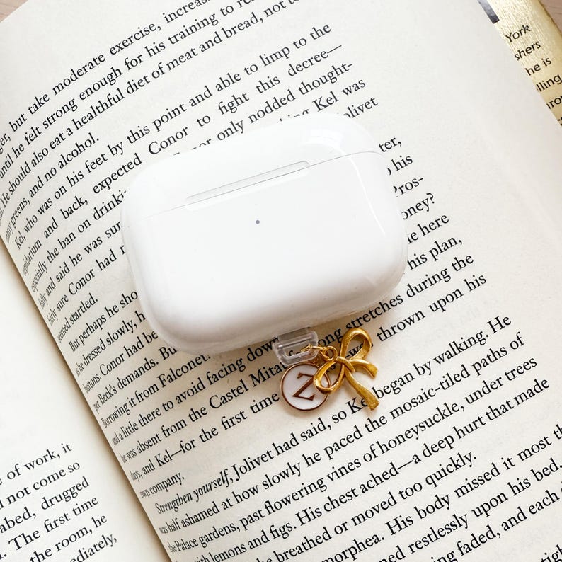 Może przedstawiać: Białe etui na AirPods ze złotym urokiem w kształcie kokardy i okrągłym urokiem z literą "Z". Etui spoczywa na otwartej książce z nadrukowanym tekstem.