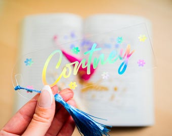 May include: A clear acrylic bookmark with the name "Courtney" in iridescent lettering and small flower accents. The bookmark has a blue tassel attached. The background is a blurred open book.