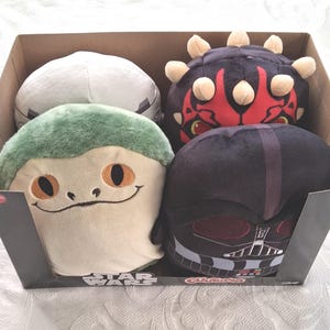 May include: A cardboard box filled with four Star Wars character plush toys. The plush toys include Darth Vader, a green creature, a white character, and Darth Maul. The box has the Star Wars logo.