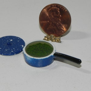 May include: A small blue and white speckled saucepan with a black handle, filled with green beads, and a matching lid. A penny is propped up nearby. This is a doll's house kitchen accessory.