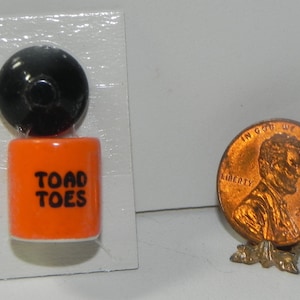 May include: A miniature orange and black ceramic container with the words "TOAD TOES" printed on the front. The container is on a white card with a black lid.