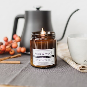 May include: A lit Cinnamon Chai scented candle in a brown glass jar with a white label that reads "wax & wool". A black kettle, a white mug, cinnamon sticks, and berries are also in the image. The candle is on a gray surface.