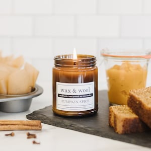May include: A lit, brown glass jar candle labeled "wax & wool PUMPKIN SPICE" sits on a black slate board with two slices of bread and a small jar of pumpkin puree. Cinnamon sticks and cloves are also on the board. A muffin tin with paper liners is in the background.