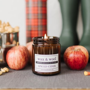 May include: A lit candle in a brown glass jar labeled "wax & wool SPICED CIDER" sits on a gray surface with apples, popcorn, green rain boots, and a red plaid thermos. Candy corn is scattered around the apples.