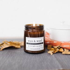 May include: A lit, brown glass jar candle labeled "wax & wool AUTUMN LEAVES" sits on a gray surface. Dried leaves and a white pot with a lid are in the background. The candle is hand-poured and made with soy wax.