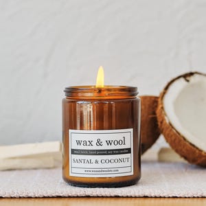 Puede incluir: Una vela encendida en un tarro de cristal marr&oacute;n, con la etiqueta "wax & wool" y "SANTAL & COCONUT". La vela est&aacute; vertida a mano y hecha con cera de soja. Se ve media coco.