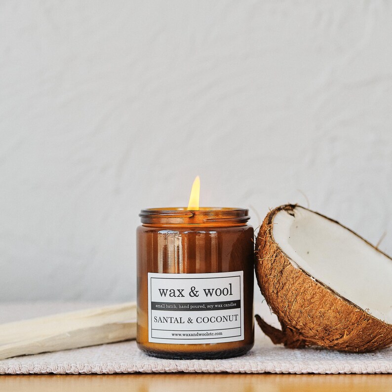 Puede incluir: Una vela encendida de color &aacute;mbar en un tarro de cristal con una etiqueta blanca que dice "wax & wool SANTAL & COCONUT". Una mitad de coco est&aacute; al lado de la vela. La vela est&aacute; sobre una superficie blanca.