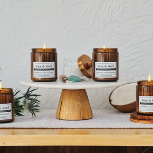 Puede incluir: Varias velas encendidas en frascos de vidrio marr&oacute;n con etiquetas blancas que dicen "wax & wool". Las velas est&aacute;n sobre un soporte para pasteles de m&aacute;rmol blanco y posavasos de madera. Los aromas incluyen Alpine Meadow, Sage & Sea Glass, Sea Salt & Sandalwood y Santal & Coconut.