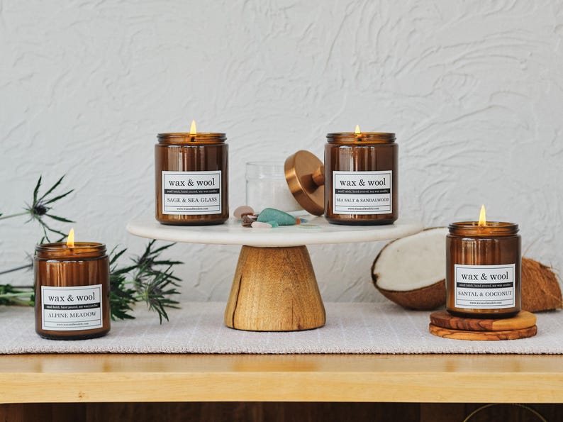 Puede incluir: Varias velas encendidas en frascos de vidrio marr&oacute;n con etiquetas blancas que dicen "wax & wool". Las velas est&aacute;n sobre un soporte para pasteles de m&aacute;rmol blanco y posavasos de madera. Los aromas incluyen Alpine Meadow, Sage & Sea Glass, Sea Salt & Sandalwood y Santal & Coconut.