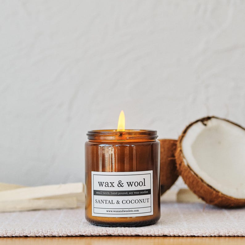 Puede incluir: Una vela encendida en un tarro de cristal marr&oacute;n, con la etiqueta "wax & wool" y "SANTAL & COCONUT". La vela est&aacute; vertida a mano y hecha con cera de soja. Se ve media coco.