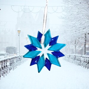 May include: A blue and teal glass snowflake ornament hanging in front of a snowy city street scene.