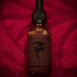 May include: A clear glass bottle with a black dropper top, filled with a brown liquid and small pieces of herbs. The label reads "Succubus oil" with a silhouette of a devil. The shop name "TheGothWitchShop" is also on the label.