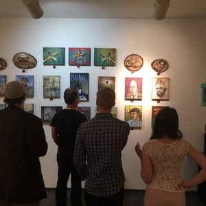 Op de afbeelding: Een galerijmuur met een verzameling diverse rechthoekige en ronde kunstwerken, waaronder schilderijen en houtbrandkunst. De tentoonstelling bevat portretten, een Batman-poster en grafische illustraties op een effen witte muur.