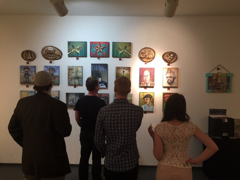 Op de afbeelding: Een galerijmuur met een verzameling diverse rechthoekige en ronde kunstwerken, waaronder schilderijen en houtbrandkunst. De tentoonstelling bevat portretten, een Batman-poster en grafische illustraties op een effen witte muur.